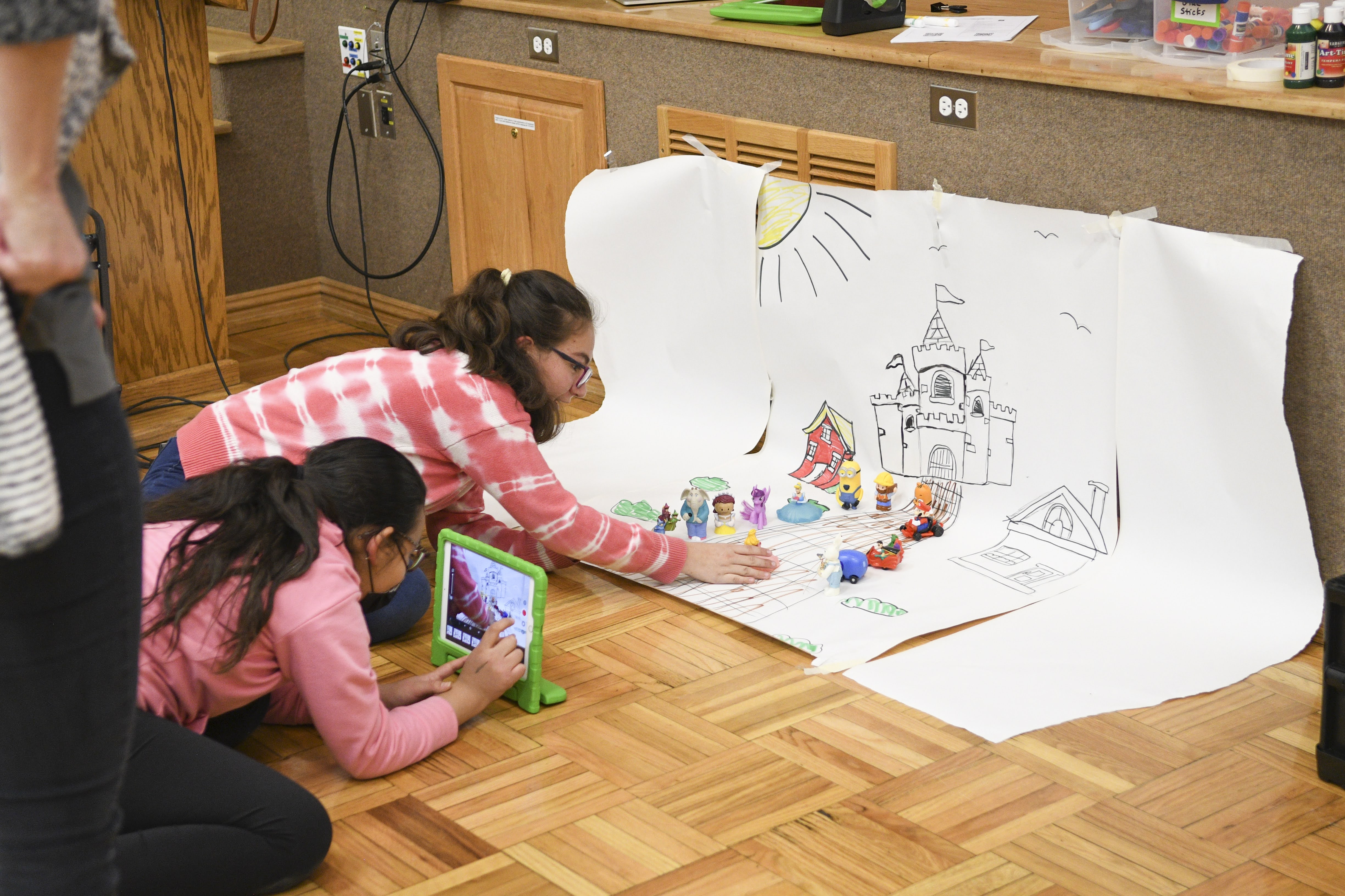 Two students on the floor filming a stop-motion scene with an iPad and hand-drawn background