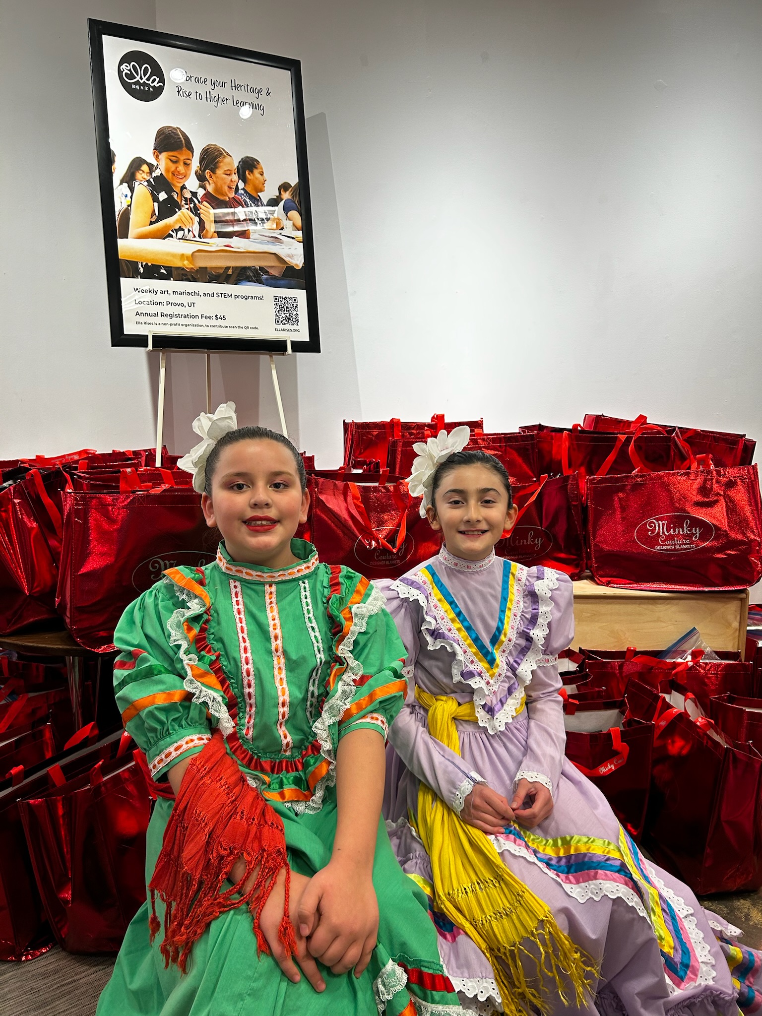Two young Ella Rises dancers in colorful folklórico dresses sitting in front of red gift bags